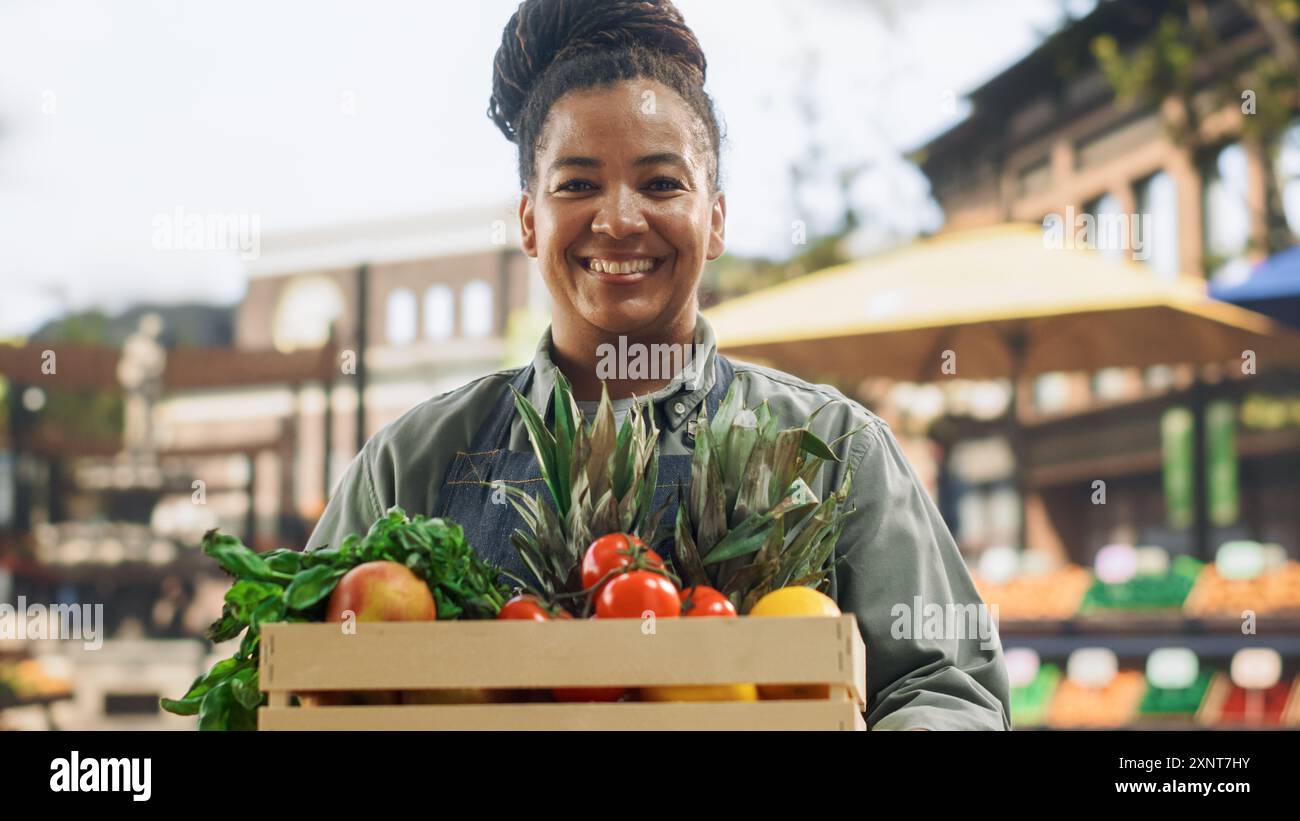 Ritratto di una donna nera che lavora in un mercato agricolo Stall with Fresh Organic Agricultural Products. Donna d'affari africana che tiene una cassa con frutta e verdura, guarda la fotocamera e sorride Foto Stock