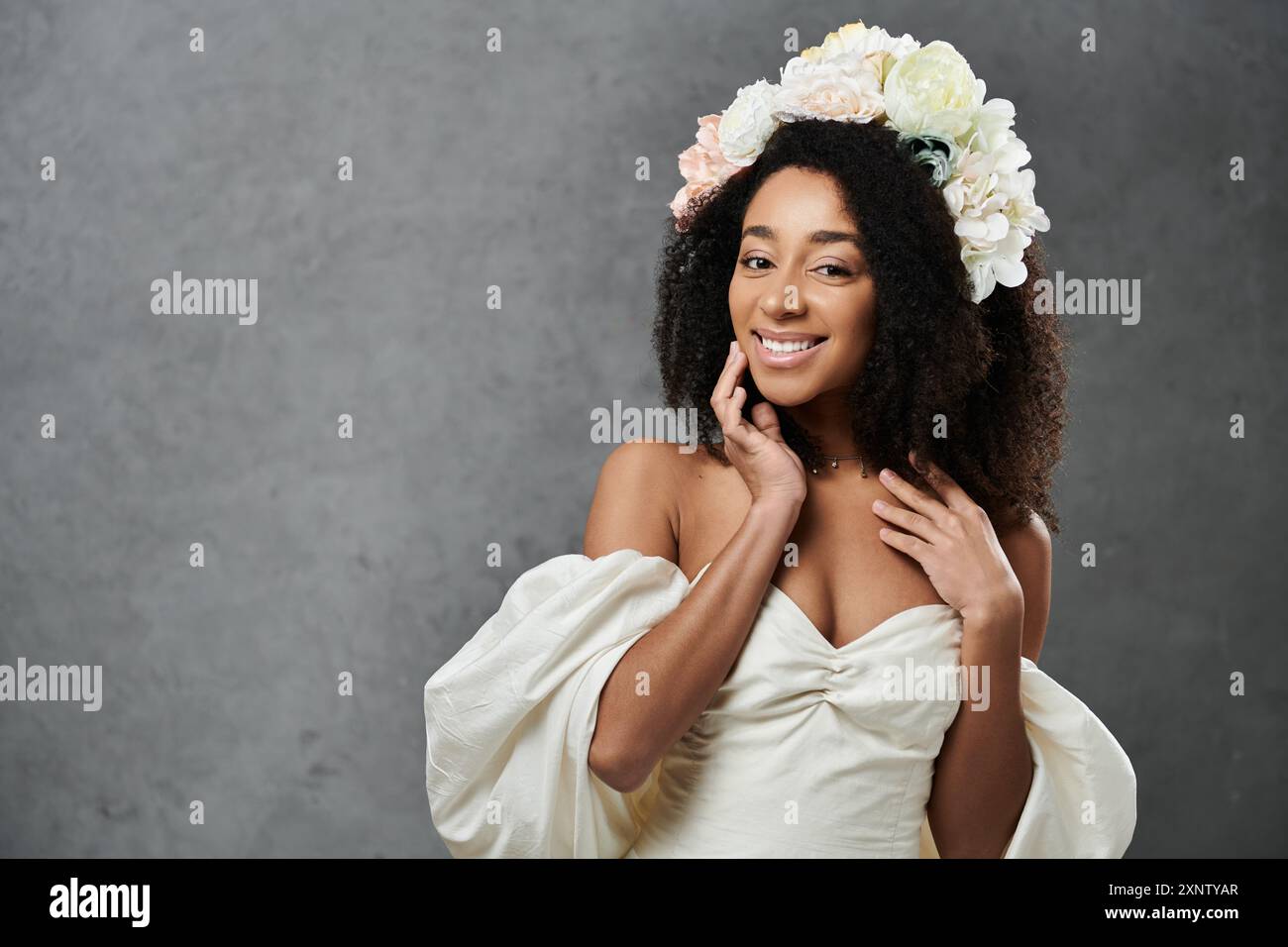 Una splendida sposa afroamericana in un abito da sposa bianco con una corona di fiori sorride radiosamente su uno sfondo grigio. Foto Stock