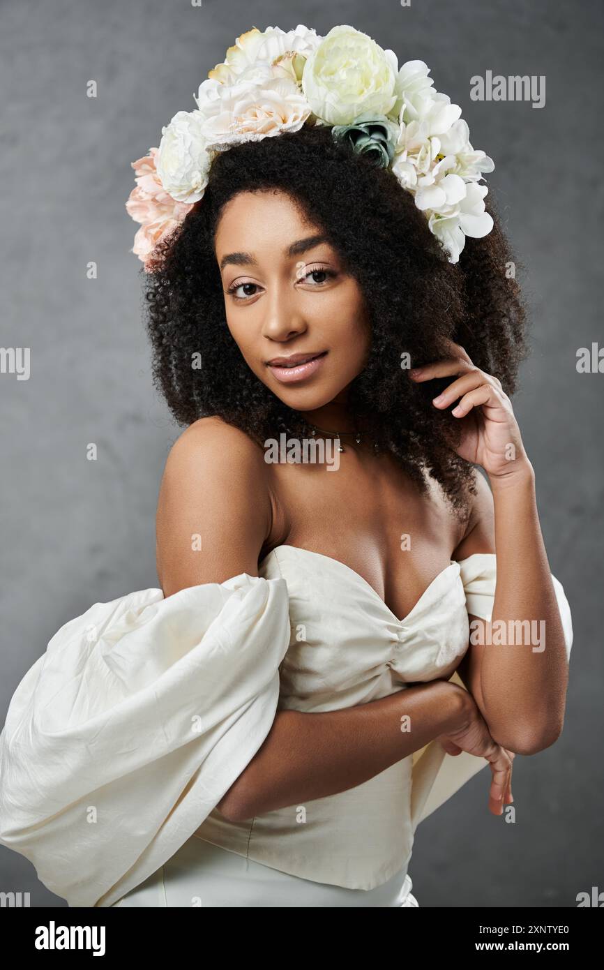 Una splendida sposa afroamericana indossa un abito da sposa bianco e una corona floreale su uno sfondo grigio. Foto Stock