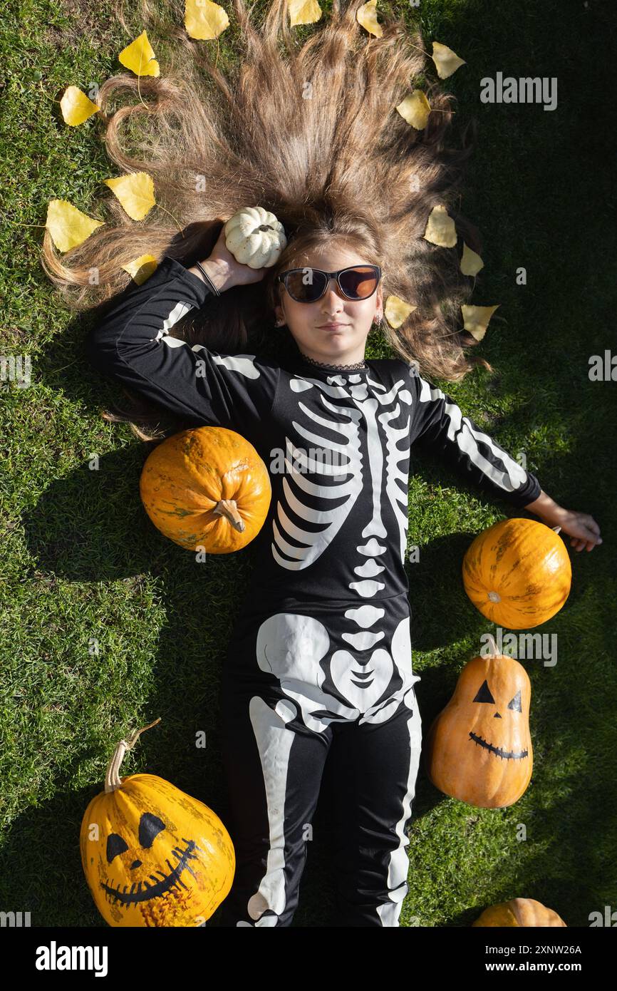 la bambina di 10-12 anni in costume nero di scheletro giace sull'erba con le zucche arancioni. Vista dall'alto. Festeggia Halloween. Bellissimi capelli con yello Foto Stock