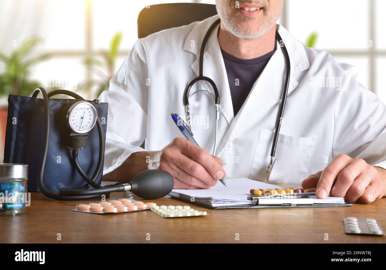 Medico che prescrive farmaci per la pressione sanguigna con diverse confezioni blister su un tavolo da ufficio in legno con misuratore della pressione sanguigna e finestra con le piante i Foto Stock