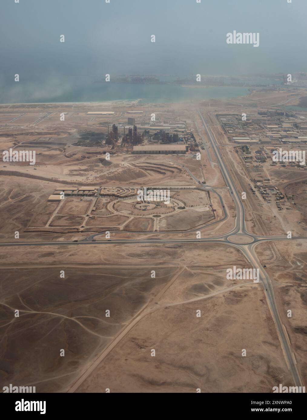 Questa fotografia aerea cattura un'area industriale circondata da un vasto paesaggio desertico in Oman, sottolineando il netto contrasto tra lo sviluppo umano e l'ambiente naturale circostante. L'orizzonte costiero aggiunge un tocco sereno al terreno accidentato. Foto Stock