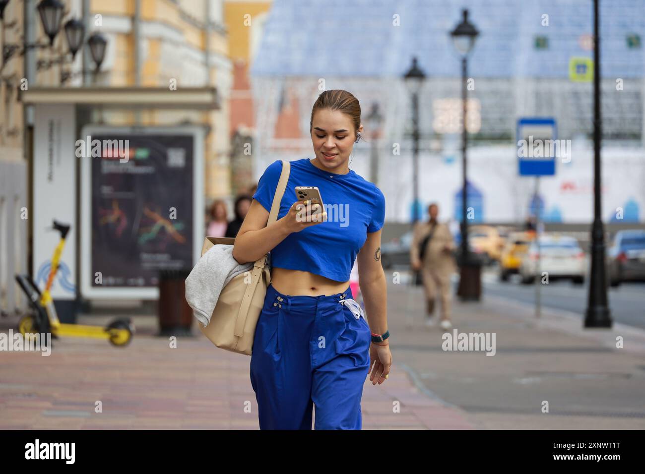 Ragazza attraente che guarda lo schermo dello smartphone che cammina per una strada. Utilizzo del telefono cellulare in città estiva Foto Stock