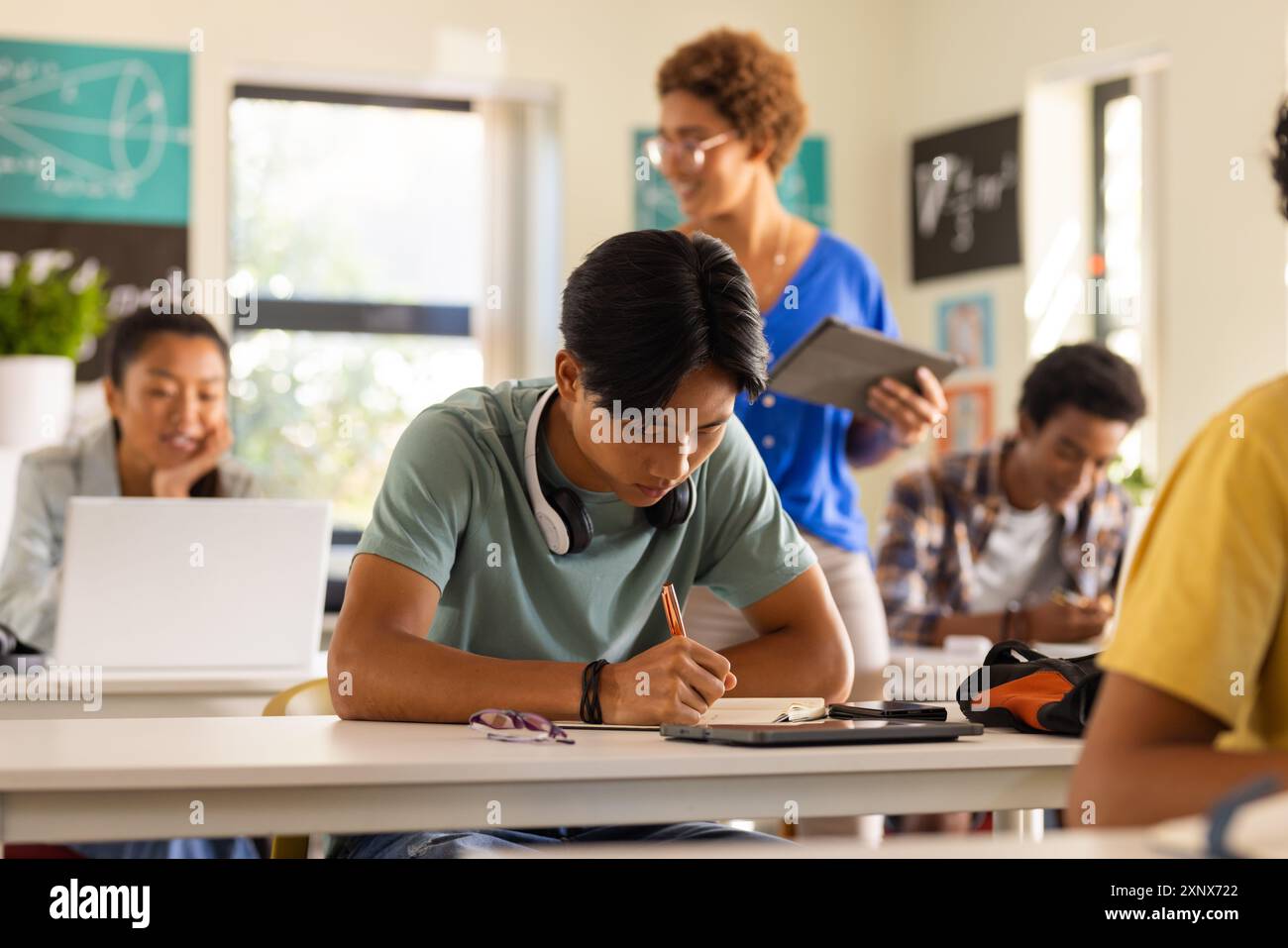 Al liceo, un ragazzo adolescente che scrive su un taccuino mentre l'insegnante con un tablet osserva Foto Stock