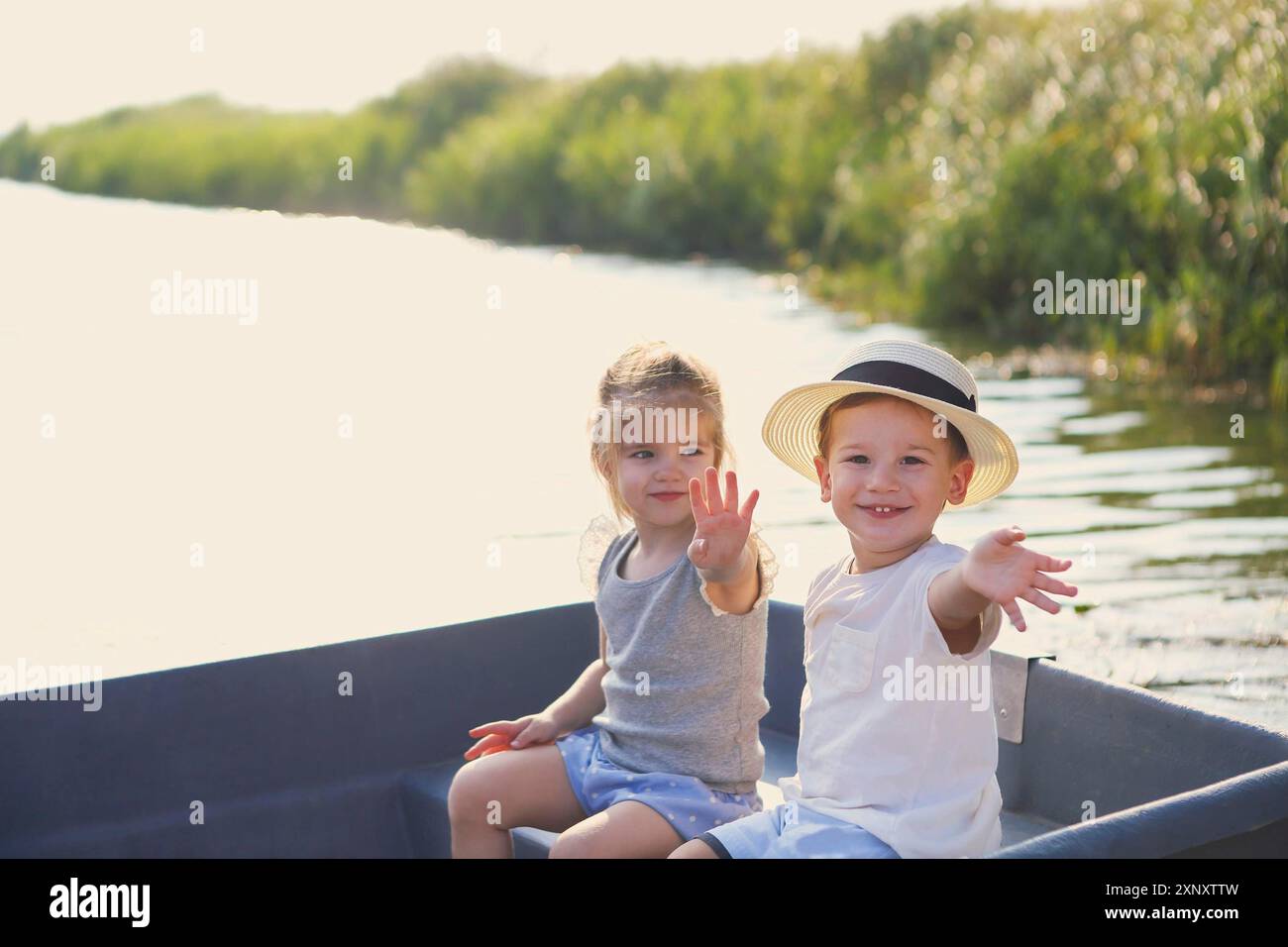 Allegro ragazzo e ragazza sorridendo e sventolando le mani mentre si siede in barca nel tranquillo fiume in campagna Foto Stock