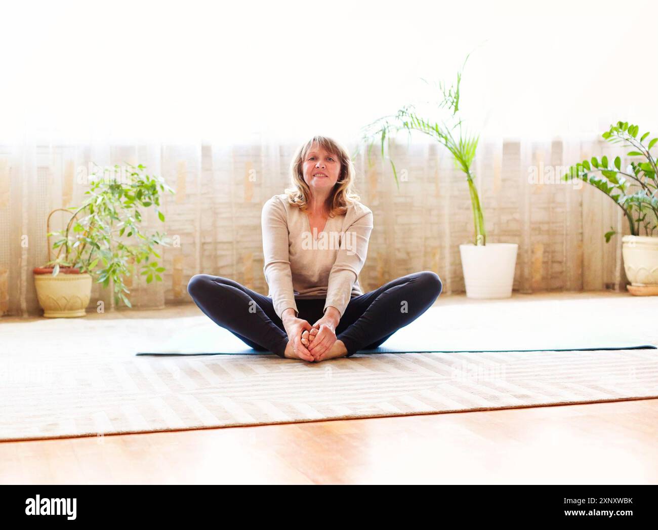 Donna anziana a piedi nudi a corpo pieno seduta sul pavimento in posizione Butterfly e gambe stretching mentre si fa yoga in camera spaziosa a casa Foto Stock