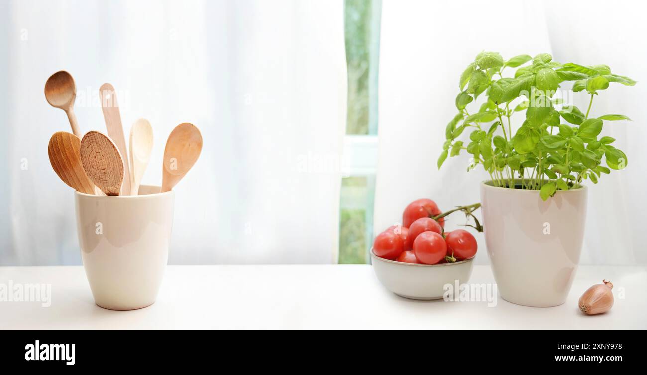 Davanzale della cucina decorato con una pianta di basilico in vaso, cucchiai da cucina in legno, ciotola con pomodori e una cipolla su tende bianche, spazio per copiare Foto Stock