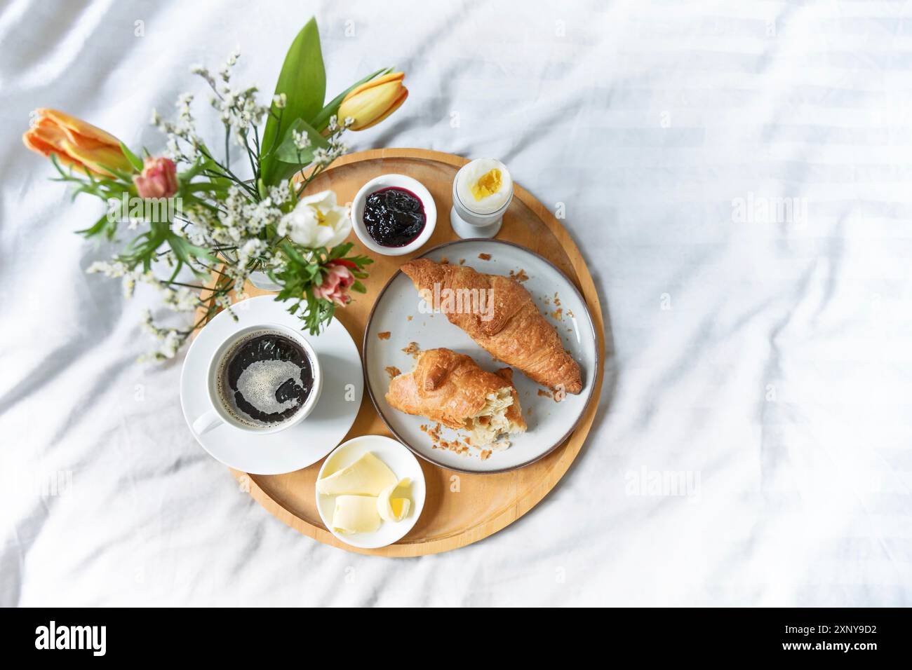 Colazione tranquilla a letto, vassoio di legno con croissant, caffè, marmellata, uova, burro e fiori su biancheria bianca verde grigia, domenica o mattina festiva Foto Stock