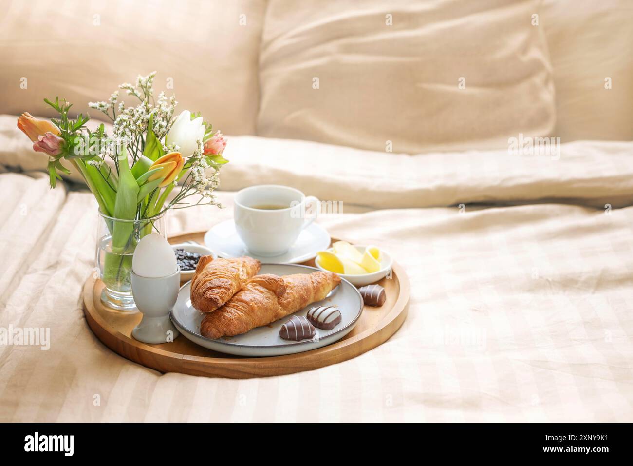 Colazione romantica a letto con fiori, croissant, tazza di caffè e cuori di cioccolato su biancheria beige per la festa delle valentine o della mamma, copia spazio Foto Stock