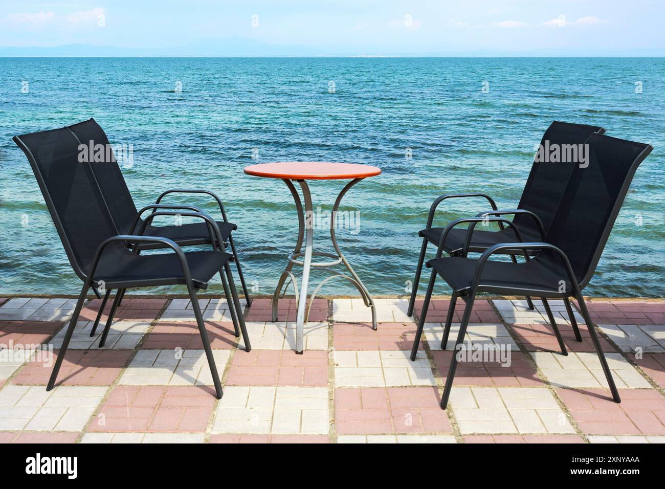 Sedie nere da esterno e un tavolo da bistrot rotondo sul lungomare lastricato direttamente sul blu del Mar Mediterraneo, invitante ambiente di relax di un Foto Stock