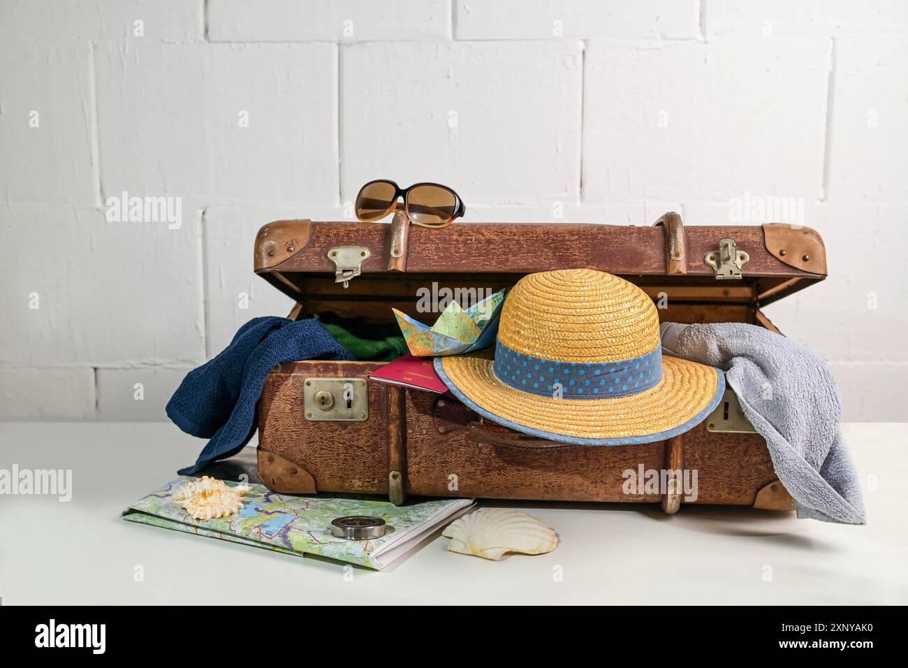 Valigia vintage con accessori da viaggio come cappello di paglia, asciugamani, passaporto, mappa e occhiali da sole per le vacanze al mare in estate, pareti bianche ruvide Foto Stock
