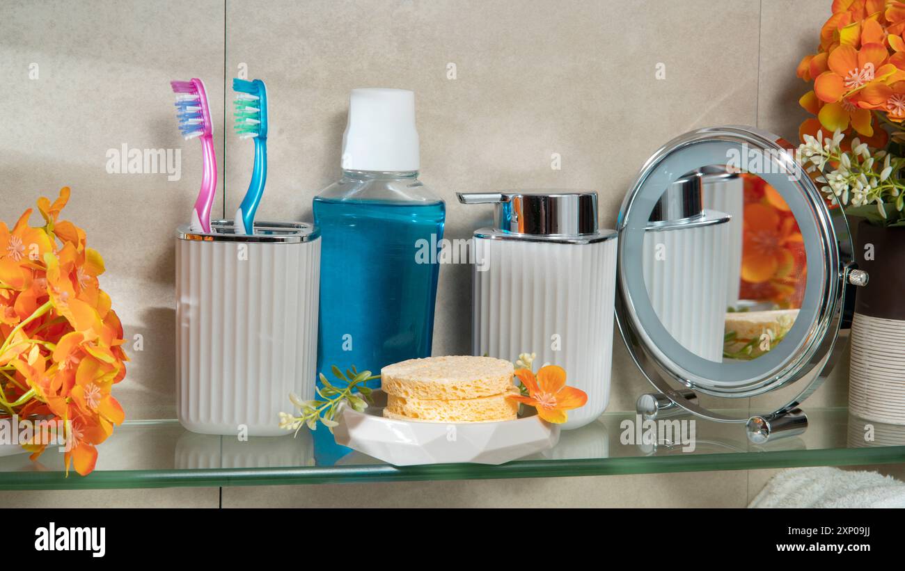 Vista ravvicinata del ripiano in vetro del bagno con bottiglia per il risciacquo vocale, spazzolino da denti, specchio, bottiglia di sapone liquido, spugne per doccia e fiori Foto Stock