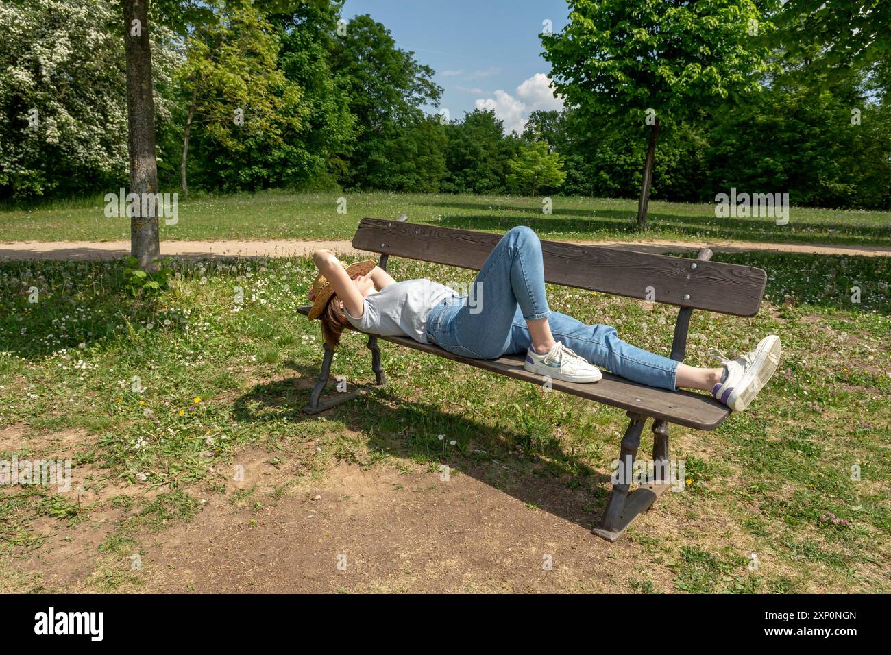 Donna con capelli marroni, t-shirt grigia, jeans che tiene il suo cappello di paglia mentre si trova su una panca parco nella natura, rilassarsi, fare un pisolino, prendere il sole Foto Stock