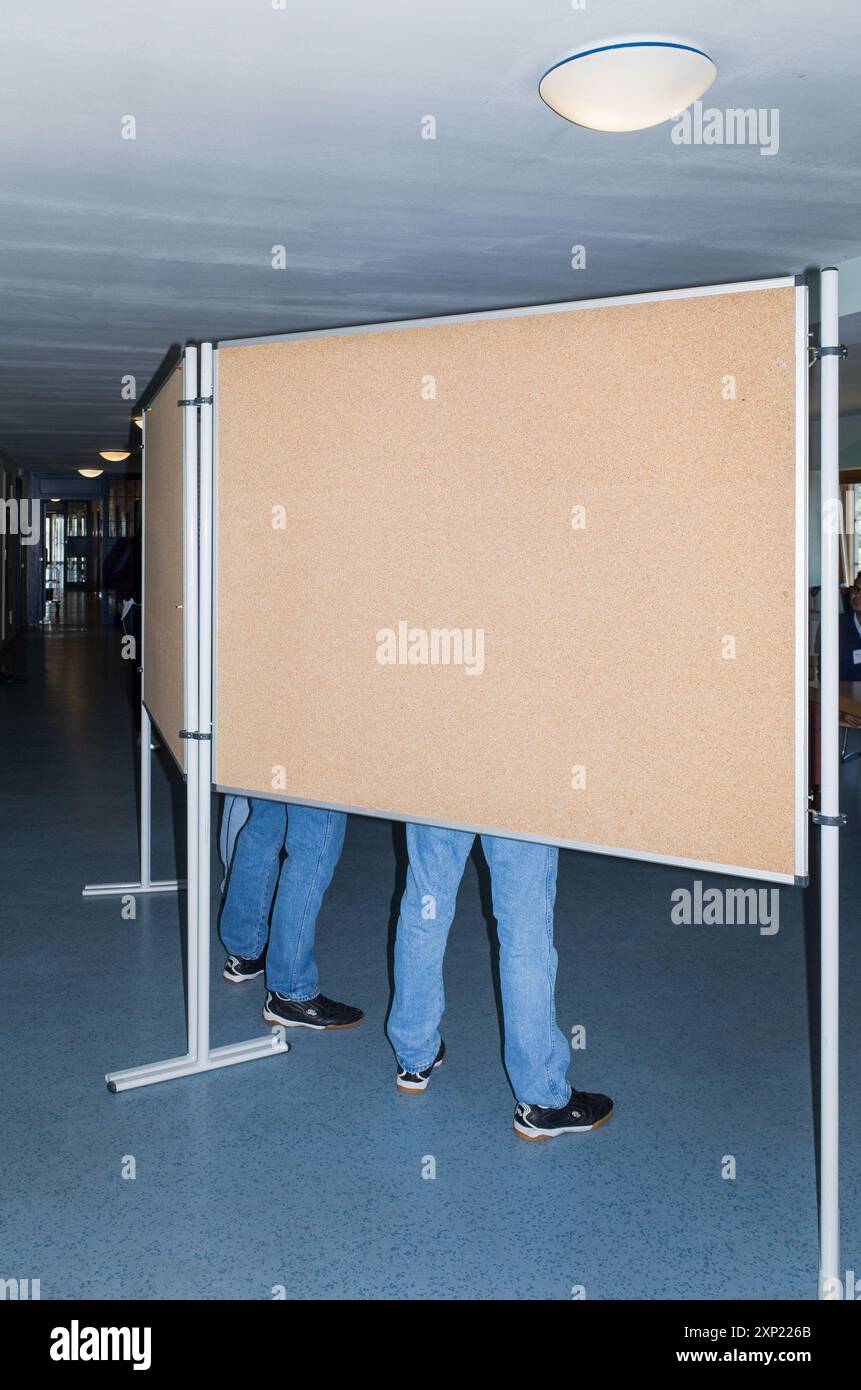 Due persone sono parzialmente visibili dietro una bacheca in un corridoio. Entrambi indossano jeans blu e scarpe casual. L'immagine evoca un senso di anonimato e curiosità. Foto Stock