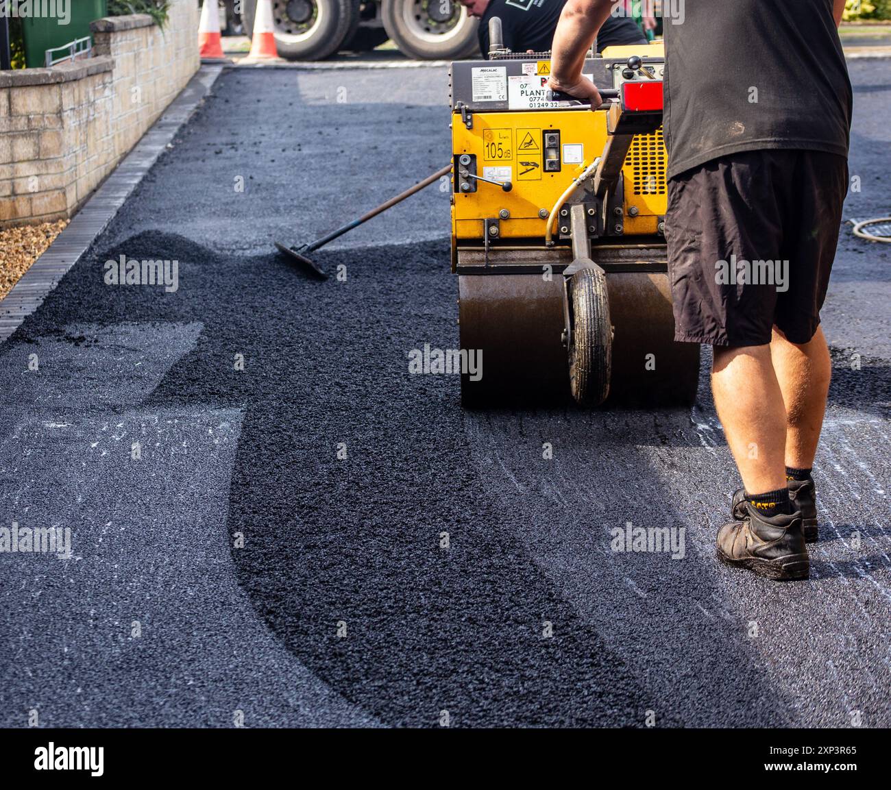 Operaio che utilizza una macchina a rulli per asfalto durante la pavimentazione di una nuova superficie del vialetto in una giornata di sole. Foto Stock