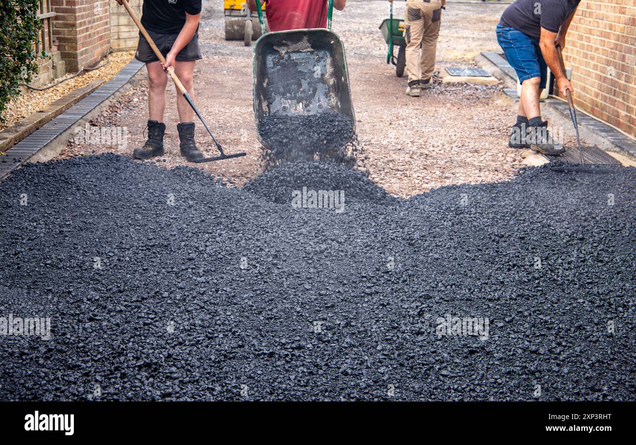 Lavoratori che utilizzano strumenti per spargere e livellare asfalto fresco su un vialetto residenziale come parte di un progetto di costruzione e pavimentazione. Foto Stock