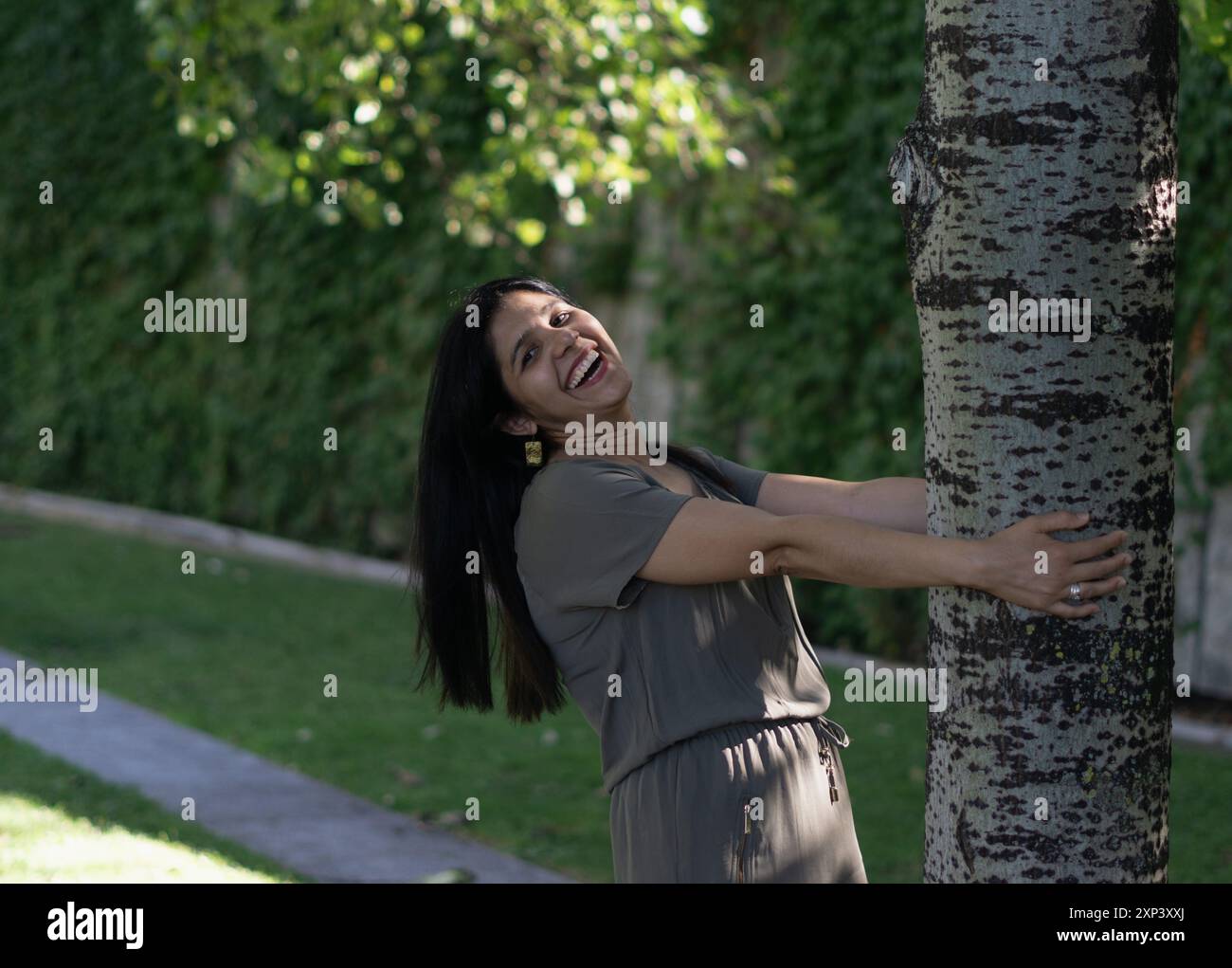 Una donna sta abbracciando un albero in un parco. Lei sorride ed è felice. Concetto di gioia e connessione con la natura Foto Stock