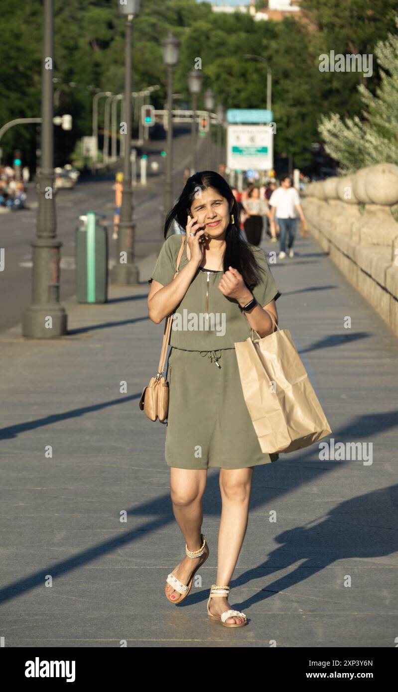 Una donna parla al cellulare mentre cammina lungo un marciapiede. Indossa un vestito verde e porta una borsa marrone Foto Stock