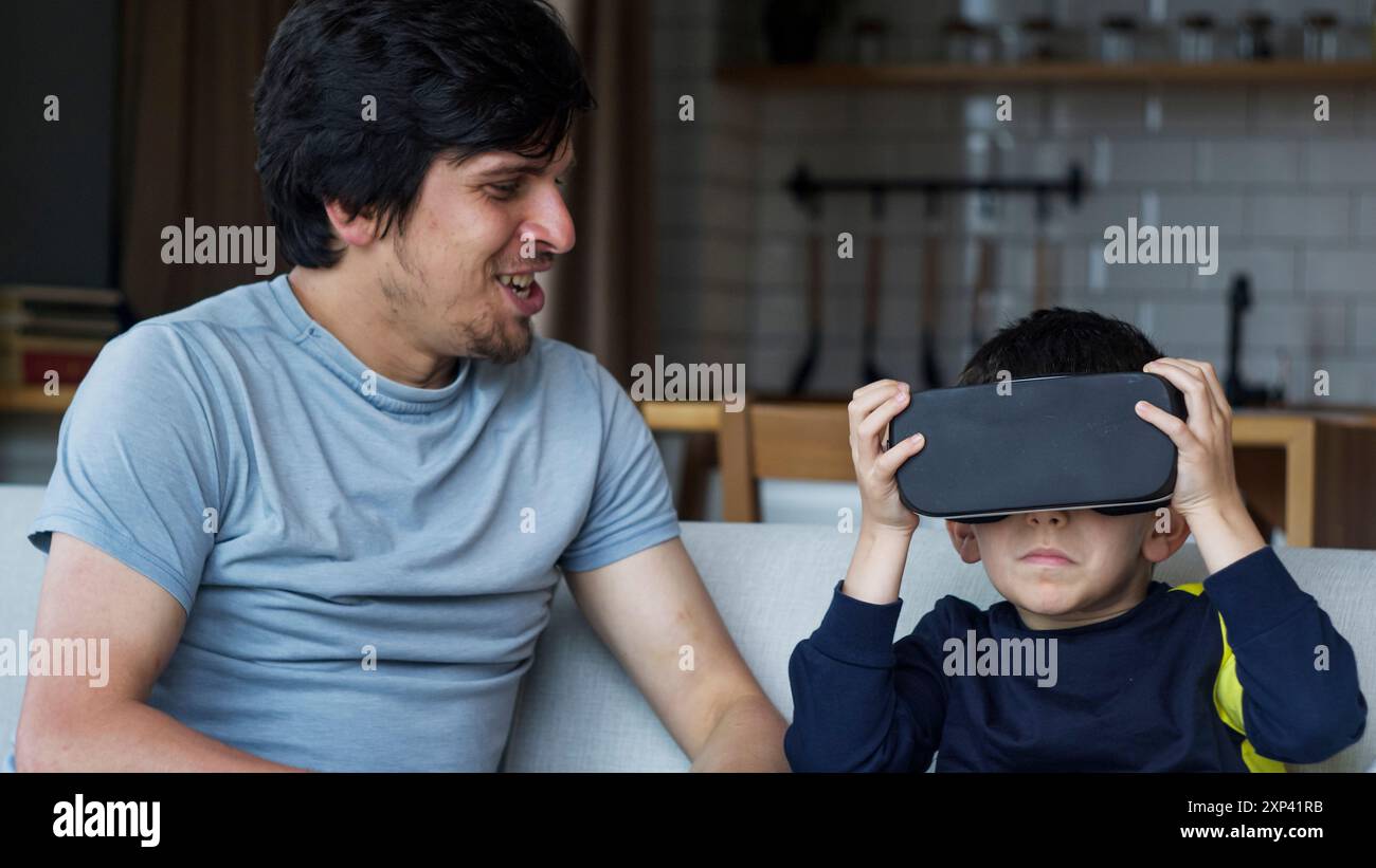 Un padre e un figlio che trascorrono del tempo di qualità a casa. Ragazzo che indossa il visore VR e sperimenta una tecnologia coinvolgente, mentre il padre guarda e sorride. Moderno Foto Stock