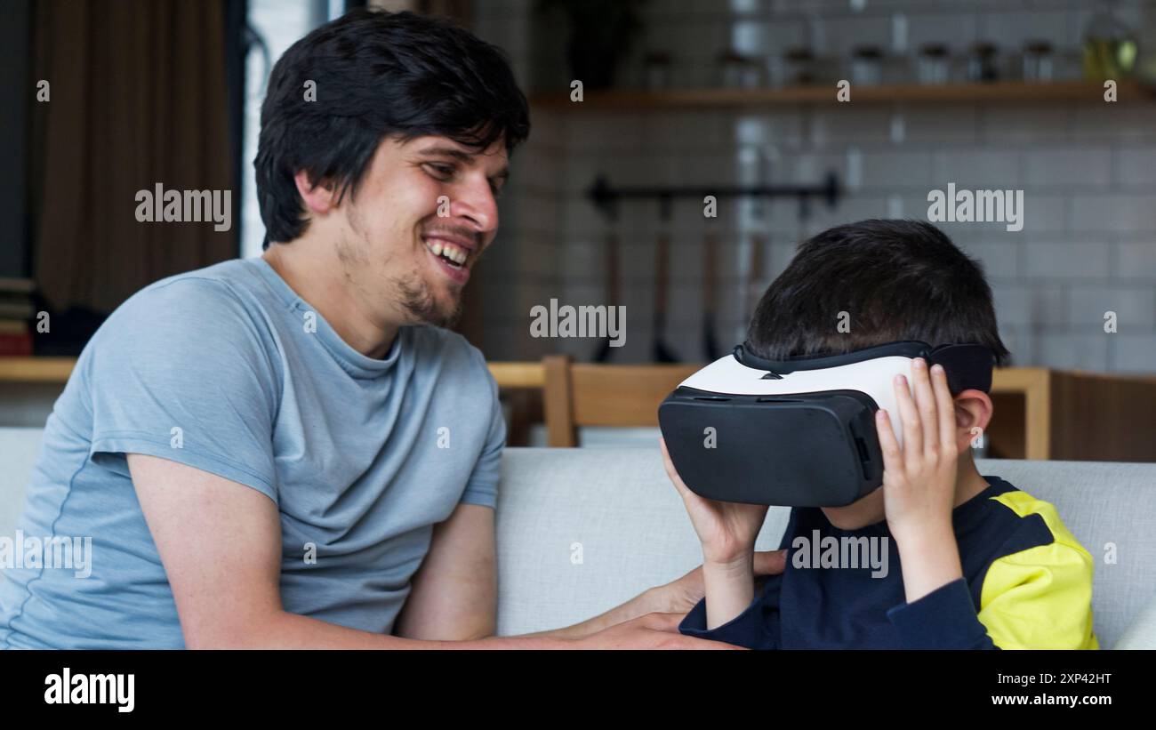 Un padre e un figlio che trascorrono del tempo di qualità a casa. Ragazzo che indossa il visore VR e sperimenta una tecnologia coinvolgente, mentre il padre guarda e sorride. Moderno Foto Stock