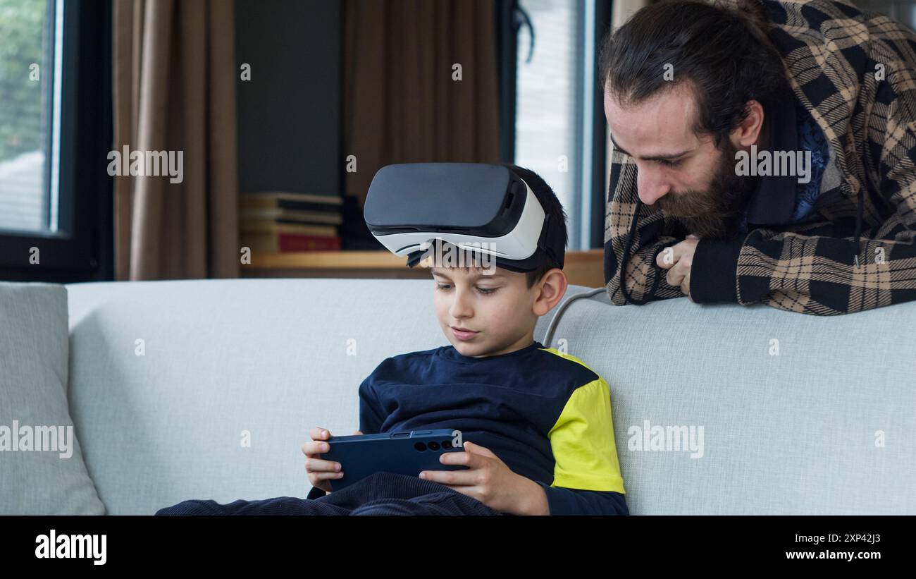 Un ragazzino seduto su un divano, indossa un visore VR e utilizza uno smartphone, mentre un uomo barbuto si appoggia per osservarlo. Tecnologia moderna e interattili familiari Foto Stock