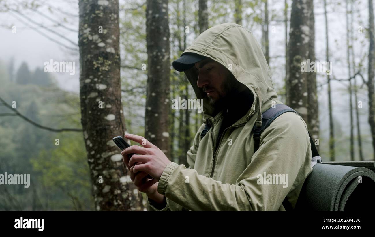 Un escursionista nella foresta usando uno smartphone, indossando una giacca con cappuccio e uno zaino. L'escursionista sta controllando il suo telefono, possibilmente per la navigazione o la comunicazione Foto Stock