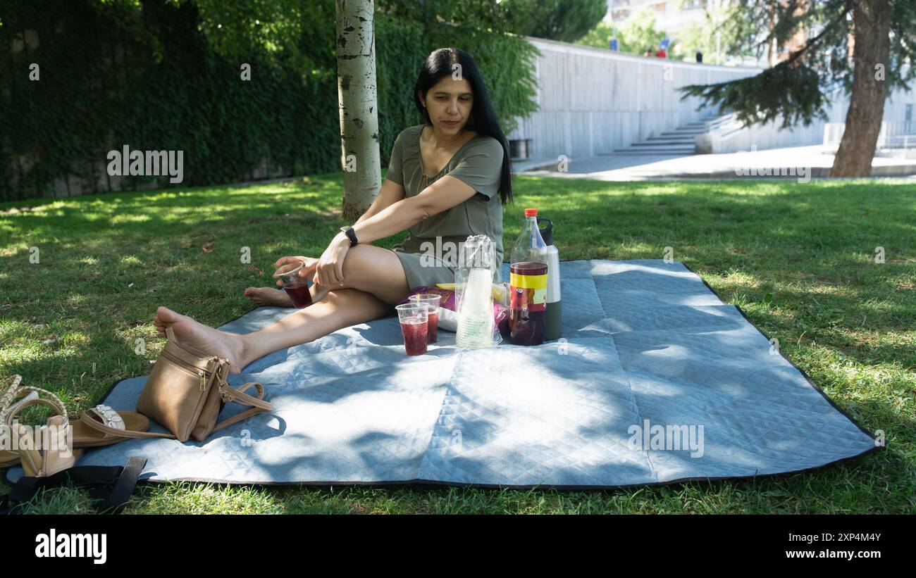 Una donna è seduta su una coperta in un parco, a godersi un picnic. Ha con sé una bottiglia d'acqua e una bottiglia di succo Foto Stock
