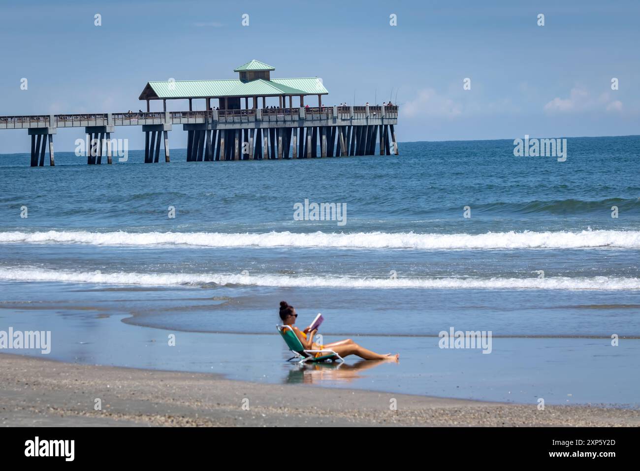Una giovane donna abbronzante e lettura Prenota in spiaggia con il molo di Folley Beach sullo sfondo Foto Stock