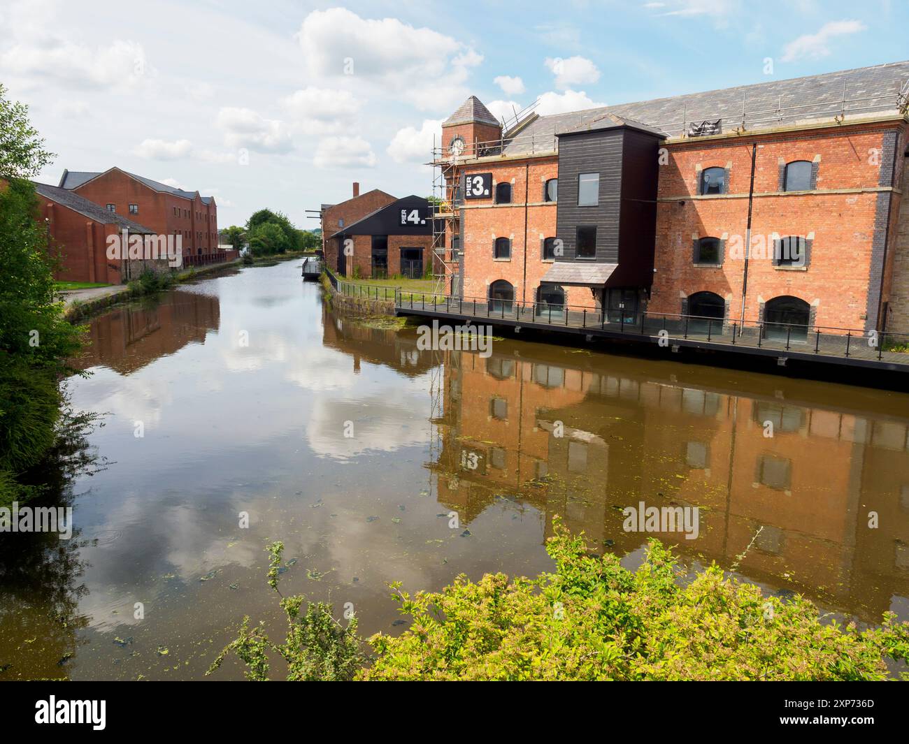 Wigan Pier Regno Unito 28 luglio 2024. Pittoresco canale con magazzini in mattoni che si riflettono sulle acque calme sotto un cielo luminoso e parzialmente nuvoloso. Foto Stock