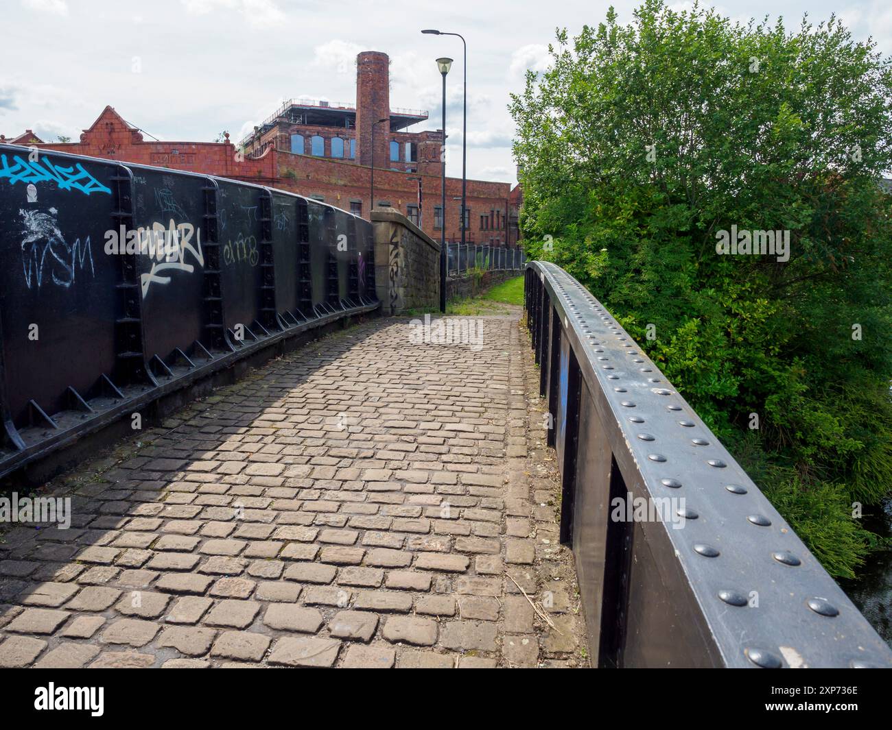 Wigan Regno Unito. 28 luglio 2024. Un sentiero acciottolato conduce su un canale a ponte in un'area urbana con pareti coperte di graffiti e vegetazione lussureggiante. Foto Stock