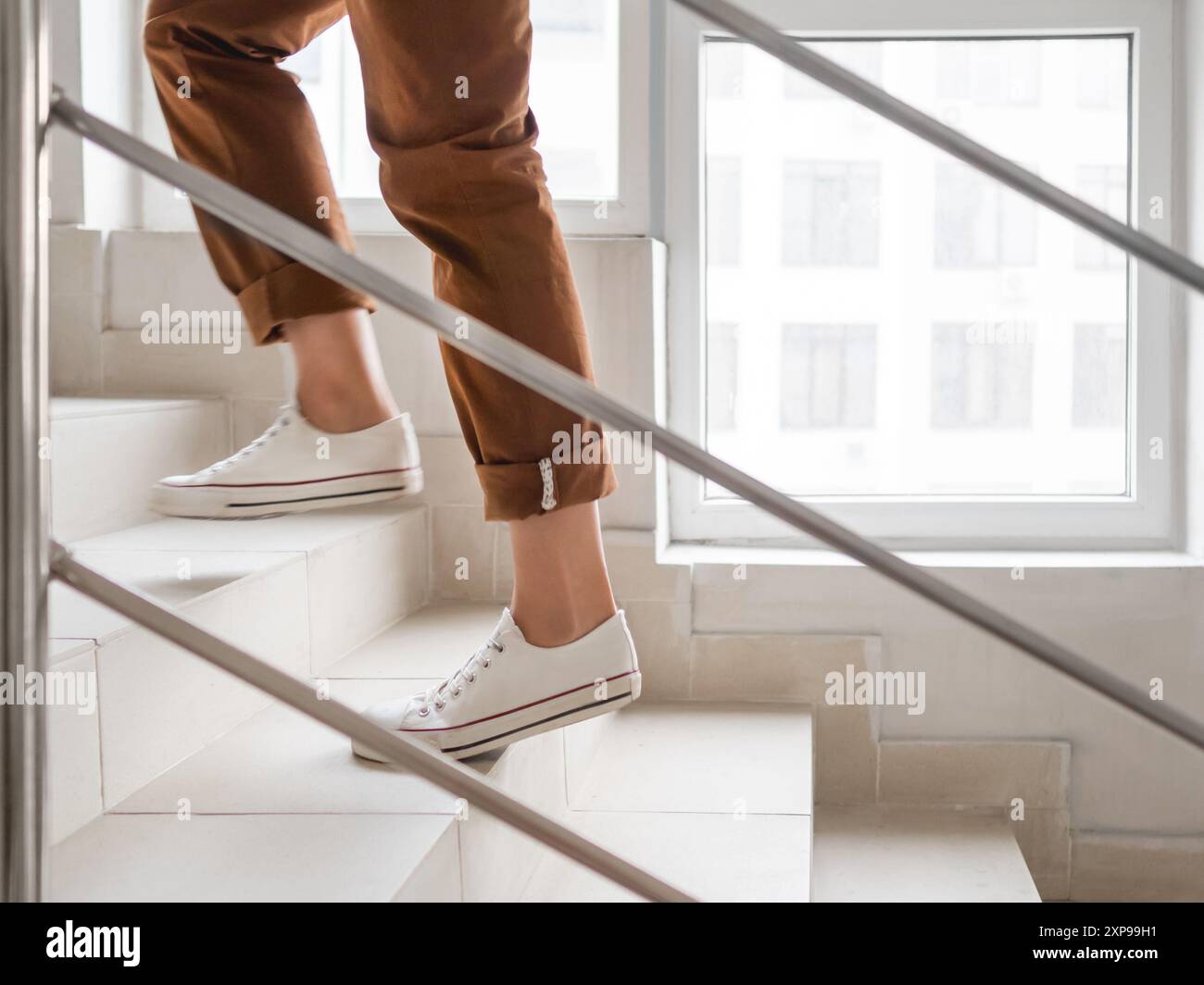 Donna in sneakers bianchi e pantaloni kaki va al piano superiore al suo appartamento. Scala bianca nell'edificio di appartamenti. Outfit casual, moda urbana. Foto Stock