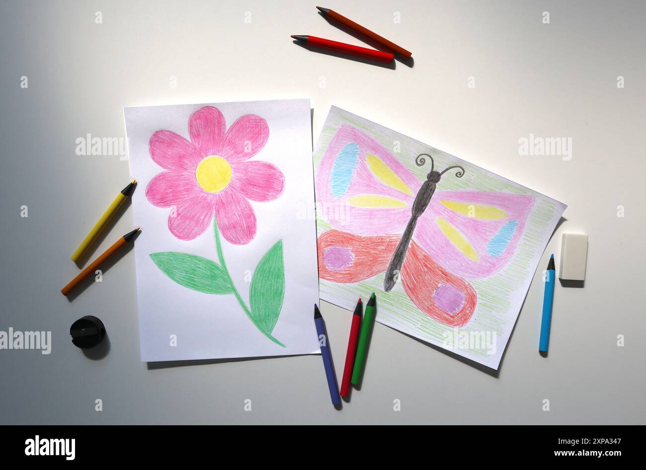 Foto di disegni per bambini con matite colorate. Uno mostra fiori, l'altro farfalla. Nelle vicinanze sono presenti matite colorate, una gomma e un affilatore. Sfondo bianco. Torna a scuola, a studiare, a fare i compiti. Foto Stock