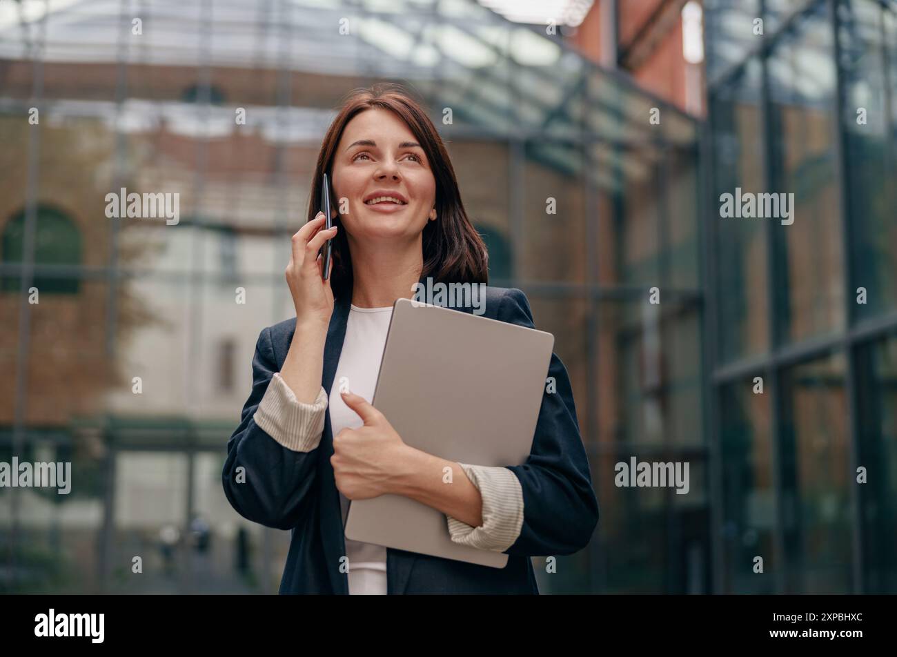 Donna d'affari sicura che utilizza lo smartphone che tiene la cartella fuori dal moderno edificio per uffici Foto Stock