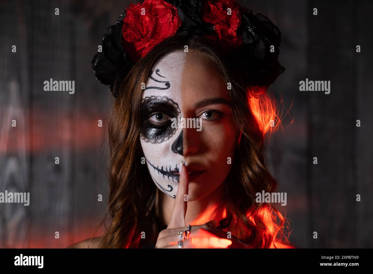 Ritratto di una donna con trucco Day of the Dead e una corona floreale, illuminata dalla luce rossa, che fa un gesto di silenzio con il dito. Foto Stock