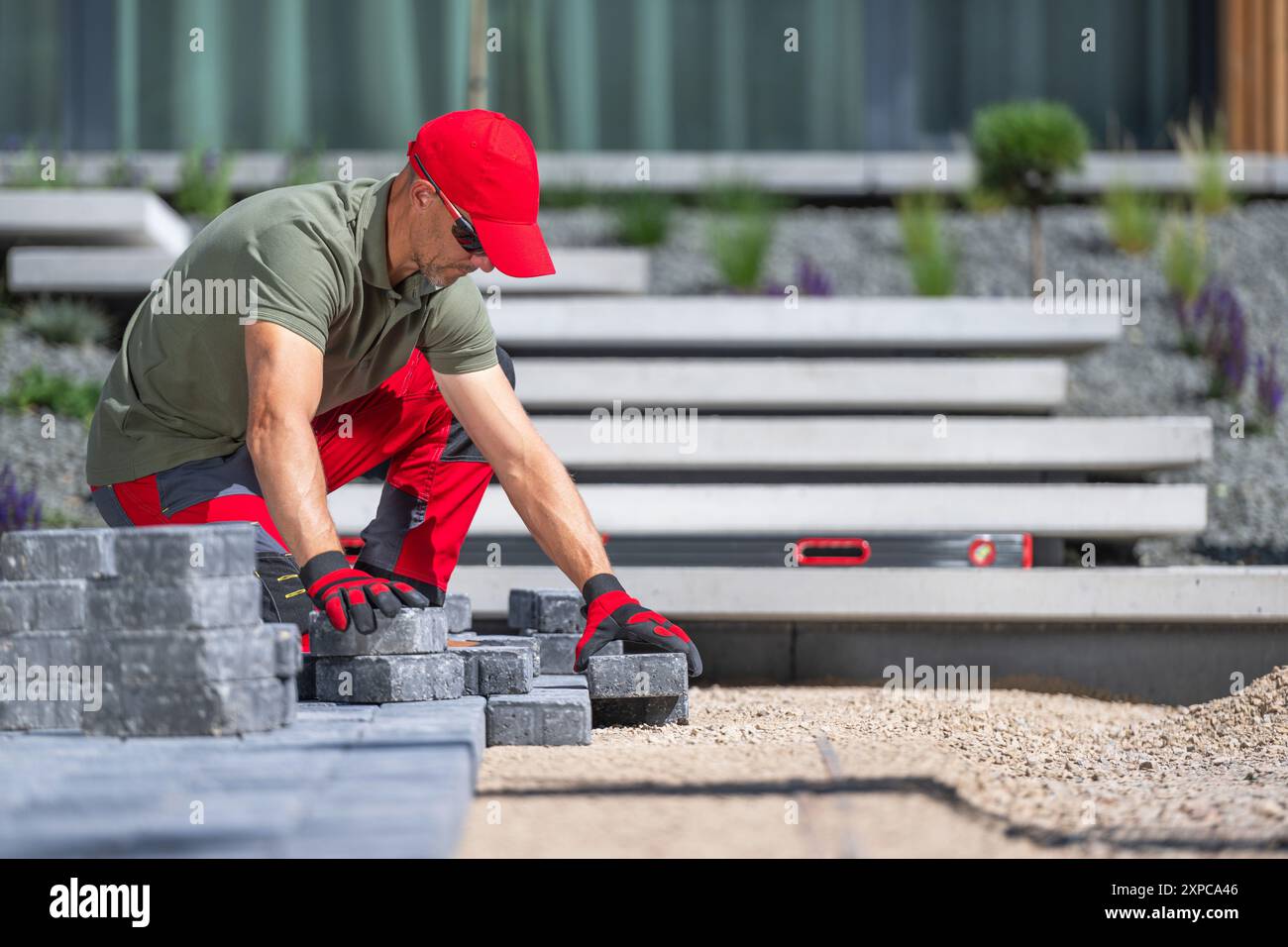 Un lavoratore sta posizionando con cura le asfaltatrici in uno spazio esterno contemporaneo indossando guanti di sicurezza e occhiali da sole. Foto Stock