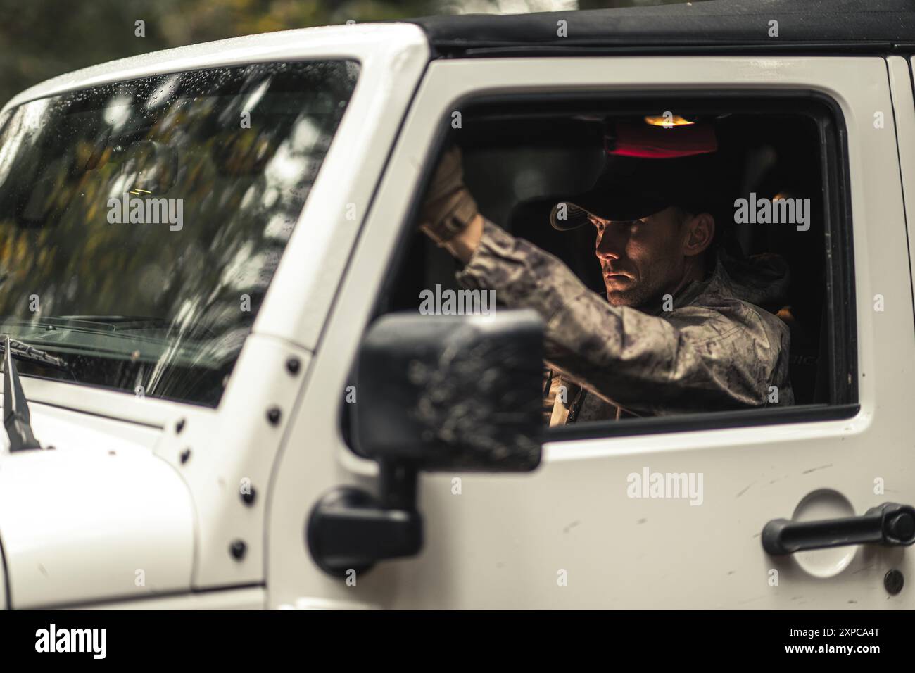 Un soldato in abbigliamento militare naviga su un SUV bianco attraverso una fitta foresta bagnata dalla pioggia, concentrandosi sul percorso che ti attende. Foto Stock