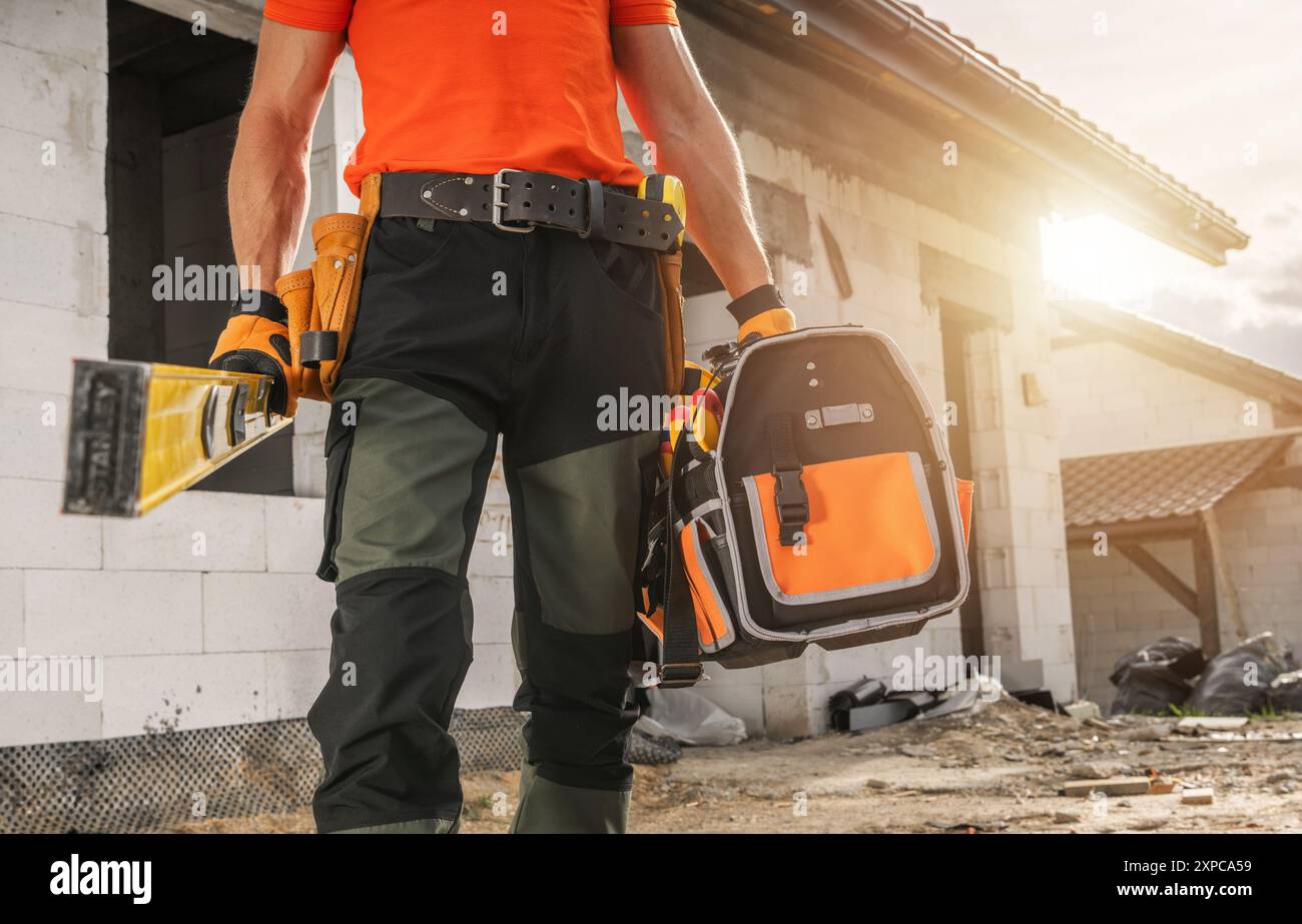 Un operaio edile si trova in un cantiere, tenendo in mano gli attrezzi e uno zaino, pronti per le attività quotidiane mentre il sole tramonta sullo sfondo Foto Stock