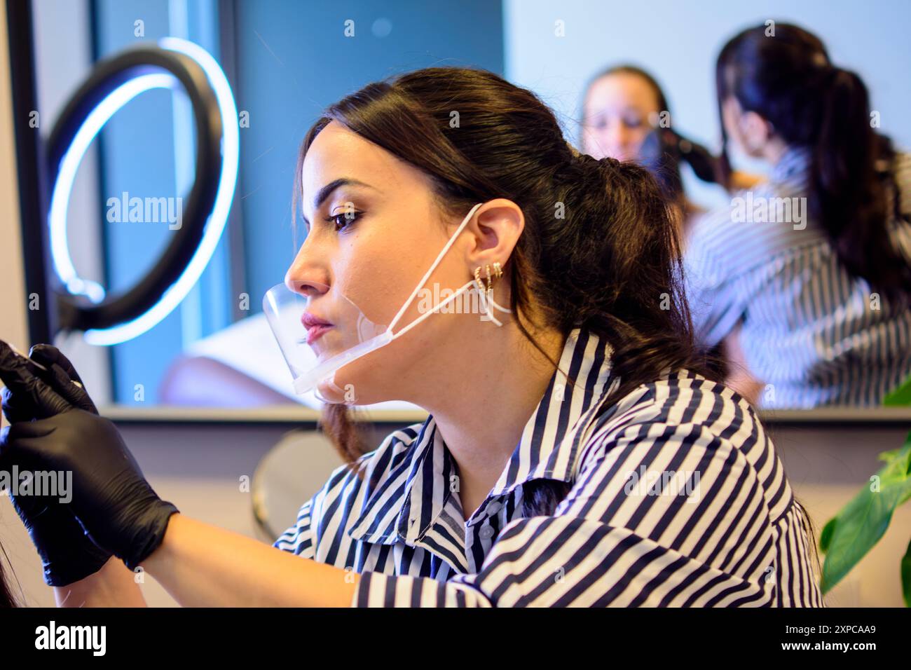 Una donna con lunghi capelli scuri, che indossa una camicia a righe e guanti, si concentra su un trattamento di bellezza in un salone. Indossa uno scudo facciale trasparente Foto Stock