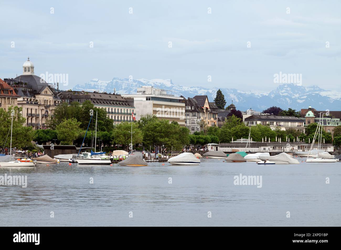 Barche e yacht parcheggiati sulla riva con edifici neoclassici e moderni e lontane cime innevate sullo sfondo Foto Stock