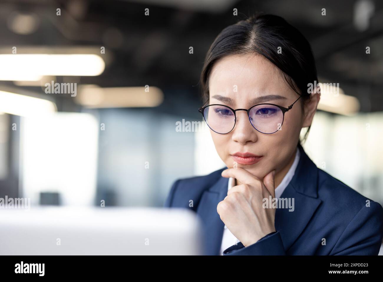 Una donna d'affari asiatica concentrata che lavora sul suo notebook in un ufficio moderno. Indossa un abito blu navy e occhiali, concentrati sui suoi compiti, mostrando determinazione e professionalità. Foto Stock