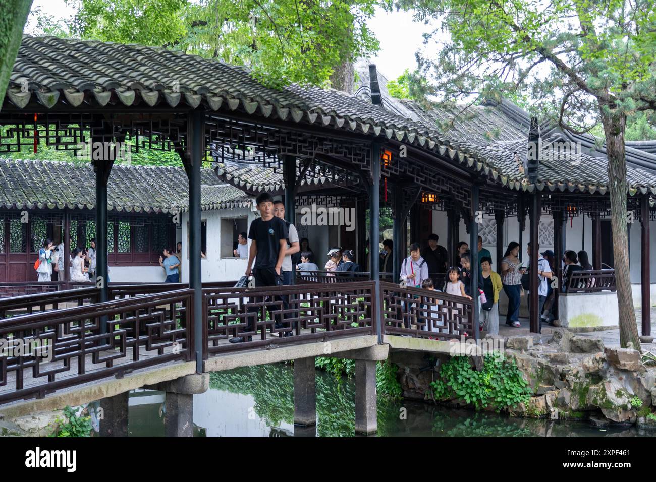 Suzhou, Cina - 11 giugno 2024: Un ponte di legno con intricate ringhiere a reticolo attraversa un tranquillo laghetto in un lussureggiante giardino cinese. Foto Stock