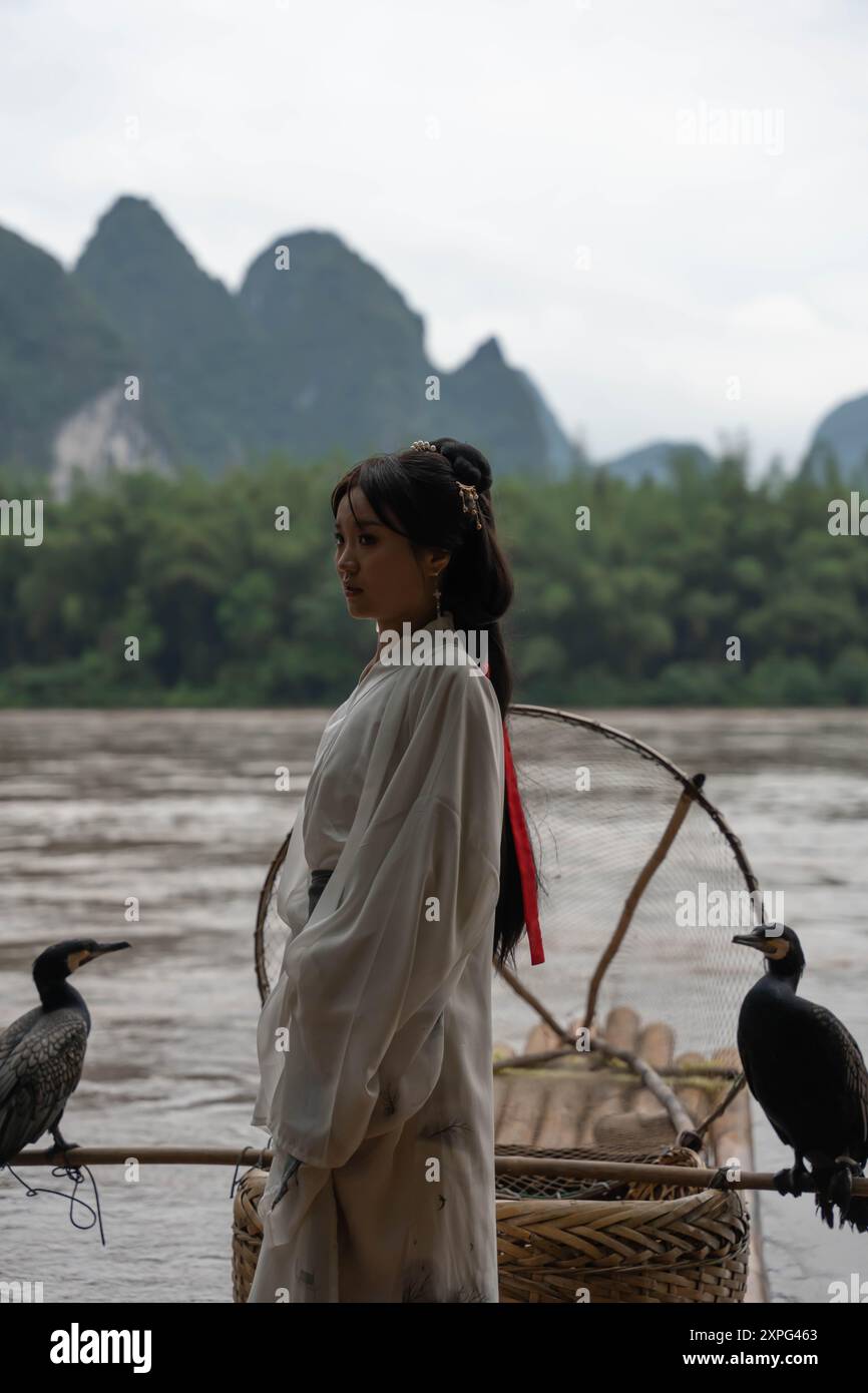 Donna di Hanfu in abito della dinastia Han su una zattera di bambù con un cormorano che fa gesti con una mano per una sessione fotografica, sullo sfondo scenico Foto Stock