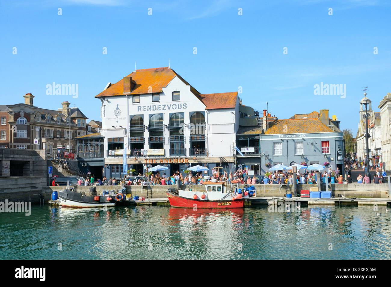 Persone che bevono al sole estivo fuori dai pub Rendezvous / The Anchor and Royal Oak, Custom House Quay, Weymouth Old Harbour, Dorset, Inghilterra Regno Unito Foto Stock