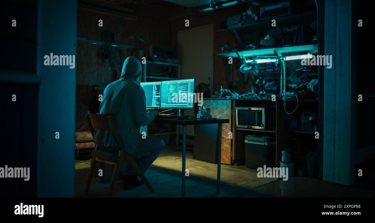 Home Garage Late at Night: Malvagio hacker con cappuccio irrompe nei server dati, attacco DDOS, attacco di phishing, malware. Uomo che scrive codice per Trojan Horse virus. Darknet Cyber Crime. Foto Stock