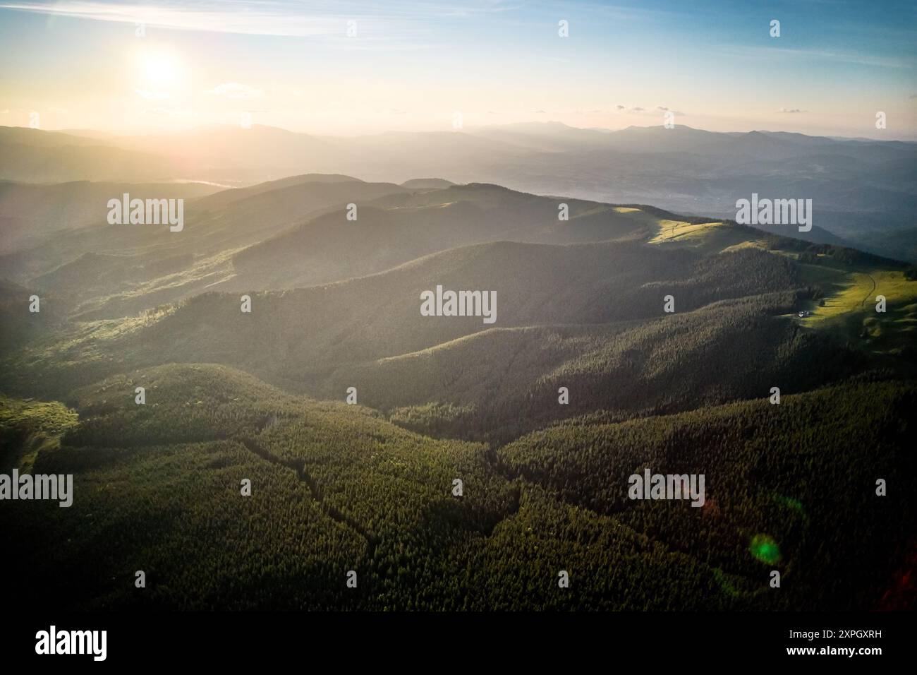 Vista aerea del tranquillo paesaggio montano al tramonto. Colline boscose immerse in una calda luce dorata, creano un'atmosfera tranquilla. Colline ondulate si estendono in lontananza, coperte da una fitta vegetazione lussureggiante. Foto Stock