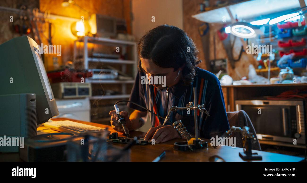 Ritratto di un ingegnere maschio caucasico che utilizza il saldatore su circuito stampato di fronte al vecchio computer desktop in un garage degli anni novanta. Studente di ingegneria che lavora al progetto per College at Home la sera Foto Stock