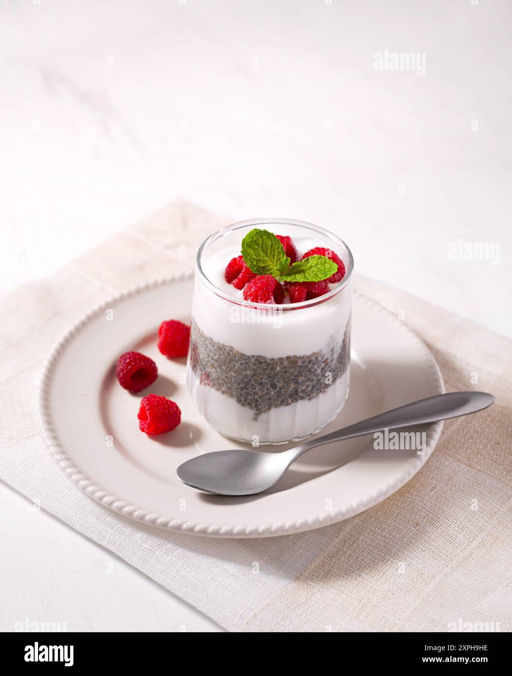 Cibo sano e crudo di yogurt al cocco, budino di chia condito con lamponi freschi maturi in un piatto bianco con un cucchiaio a lato Foto Stock