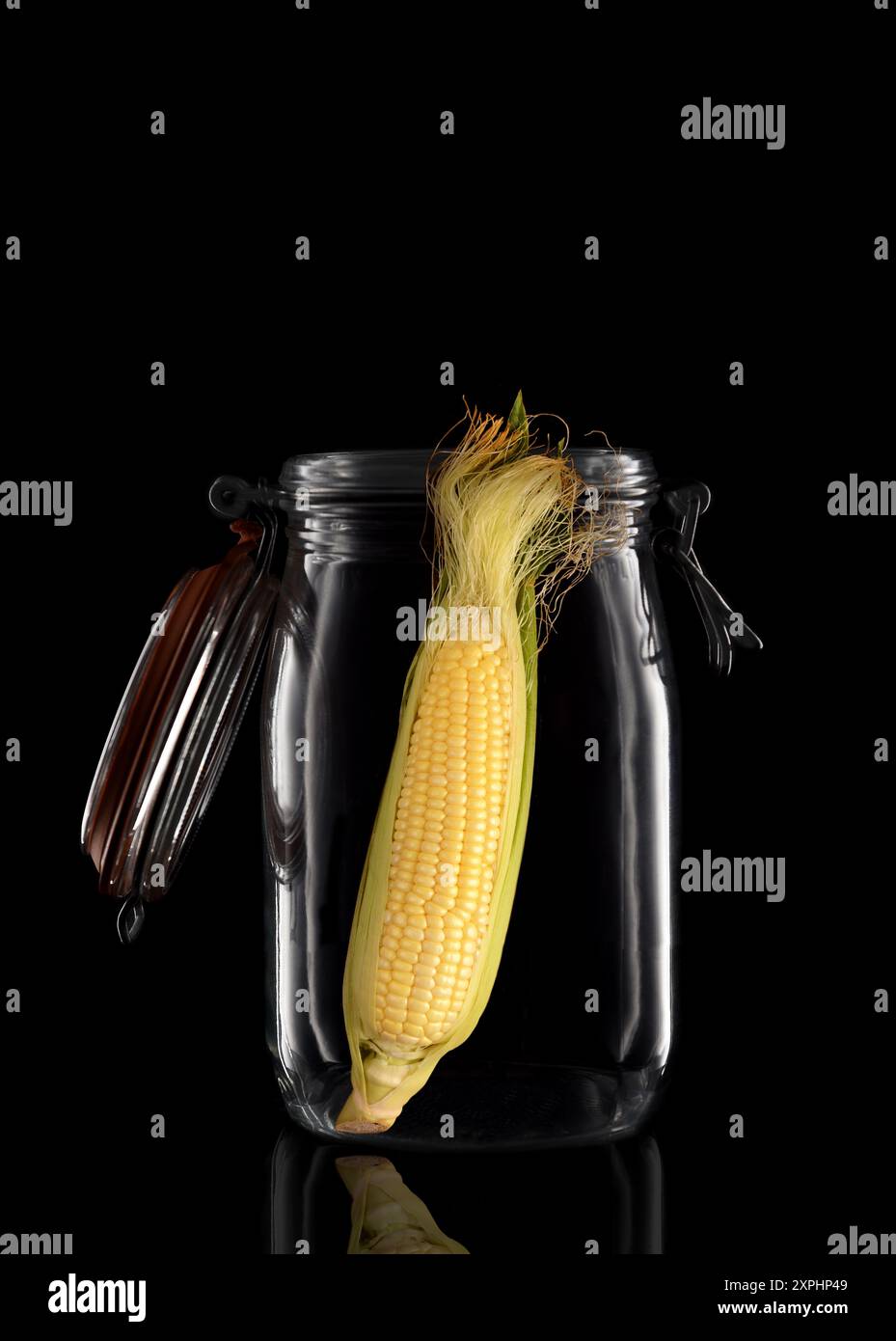 Un orecchio di granturco in una vaschetta di conservazione in vetro o in una vaschetta isolata sul nero con riflesso, con coperchio aperto. Foto Stock