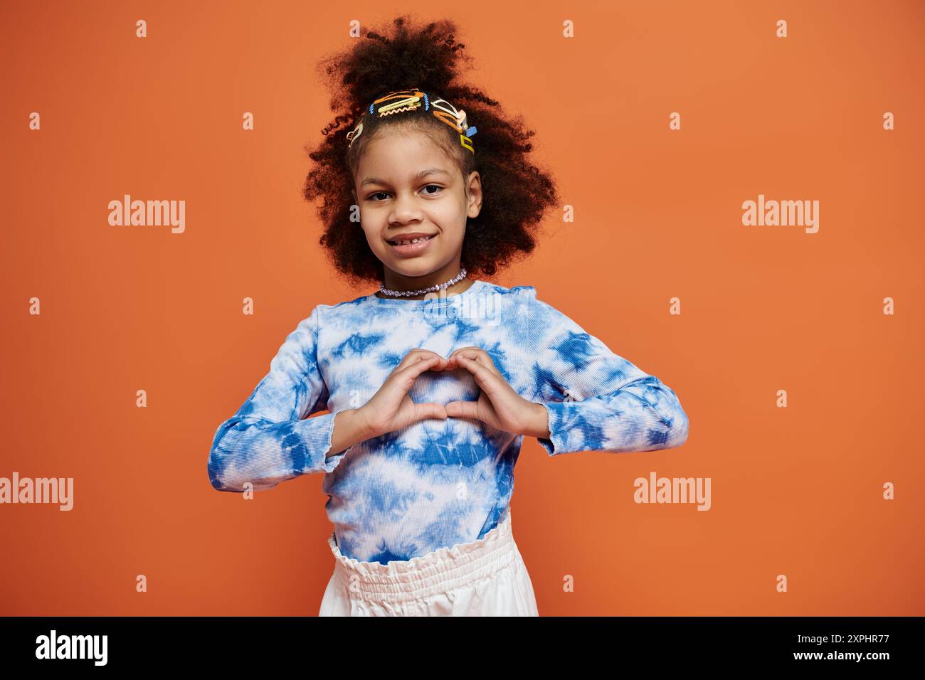 Una giovane ragazza afroamericana in un abito alla moda si erge su uno sfondo arancione vibrante, sorridente e modellando il cuore con le sue mani. Foto Stock