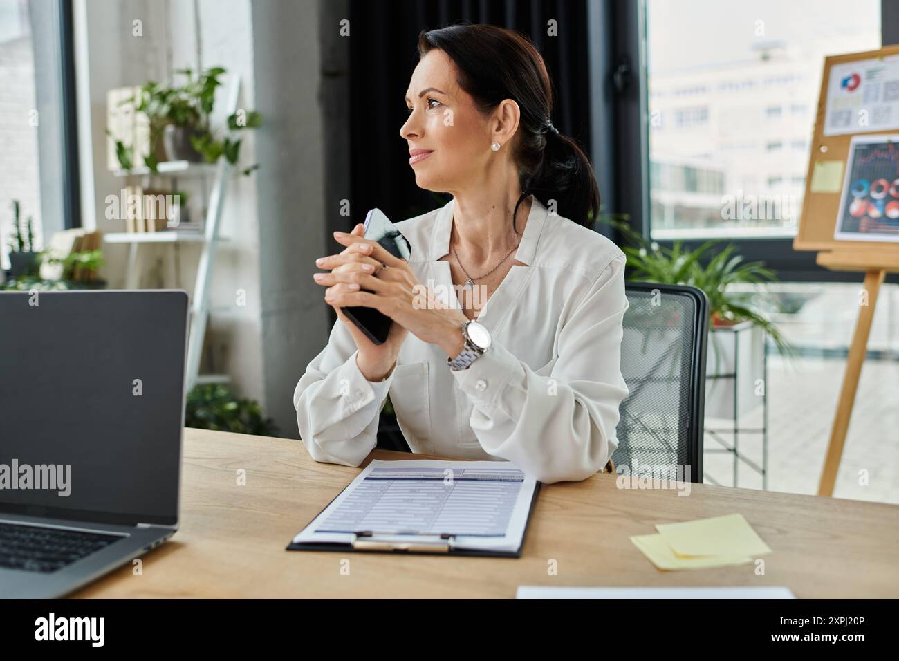 Una donna con una camicia bianca siede a una scrivania in un ufficio, regge un telefono e sembra premurosa. Foto Stock